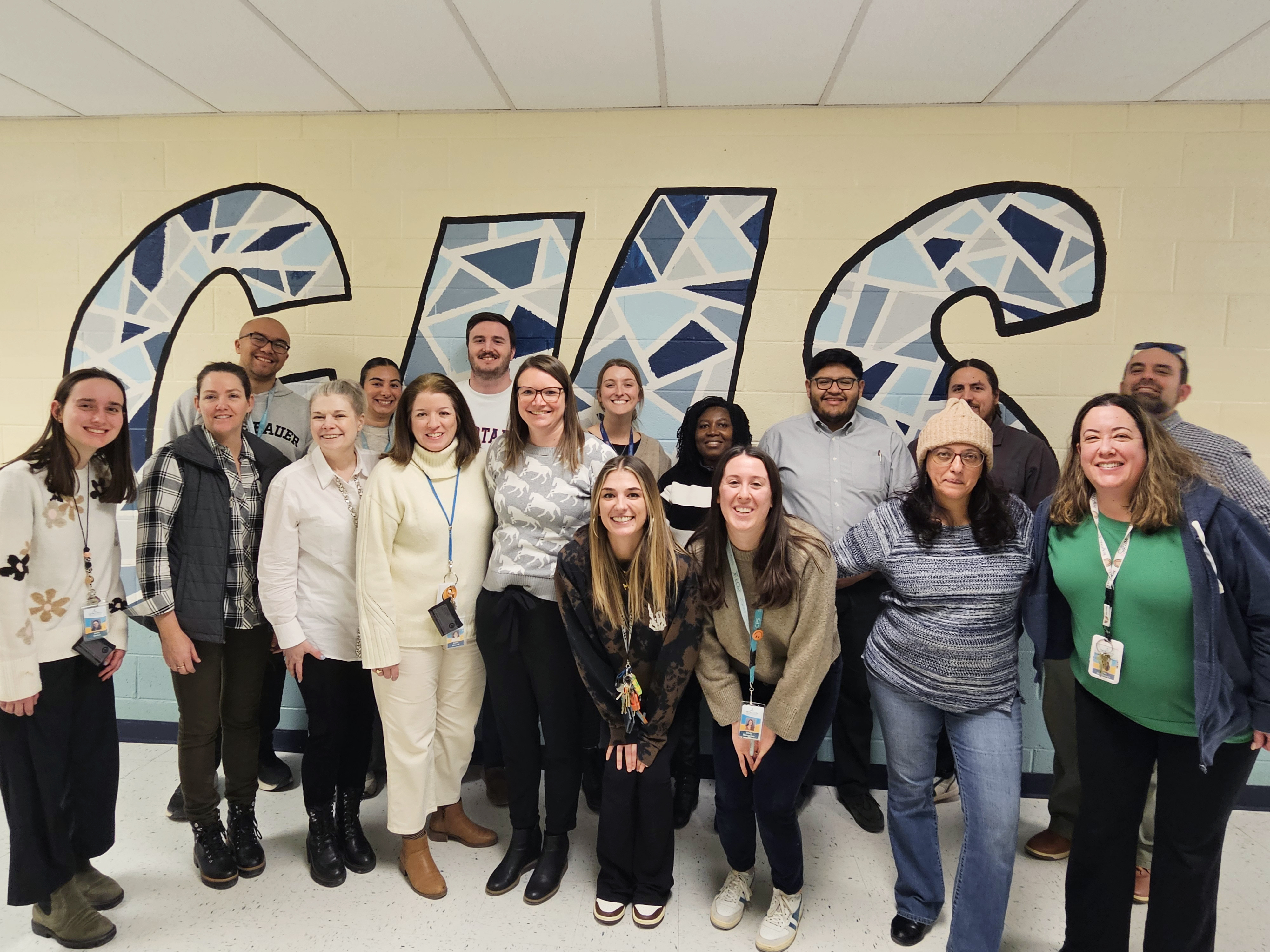 A larger group of adults pose for a group photo in front of three large blue letters: "GHS."